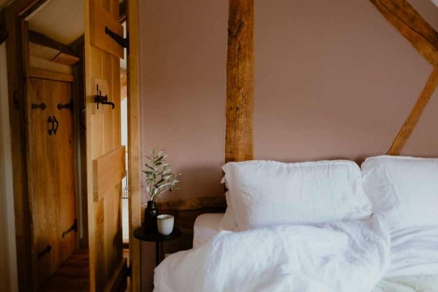 Corner of an attic bedroom with a dusky pink wall and exposed oak beams, white linen bedding, and a small side table holding a mug and an olive sprig, beside a rustic plank door with iron latches.