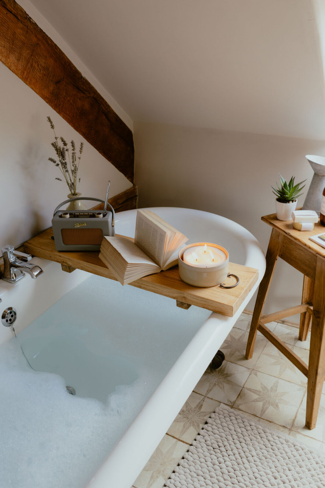 Close-up of a bubble-filled roll-top bath with a wooden bath tray holding a vintage radio, open book and lit candle, beside a rustic side table with a succulent and soaps on star-patterned tiles.