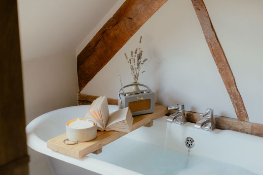 Claw-foot bath under sloping ceilings and oak beams, filling with water from chrome taps, with a candle, open book, radio and dried lavender on a wooden bath tray.