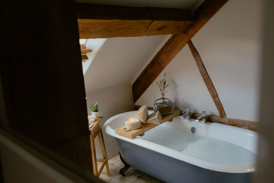 Glimpse through a doorway into an attic bathroom with oak beams, where a claw-foot bath fills beneath a wooden tray holding a candle, open book, radio and dried lavender.