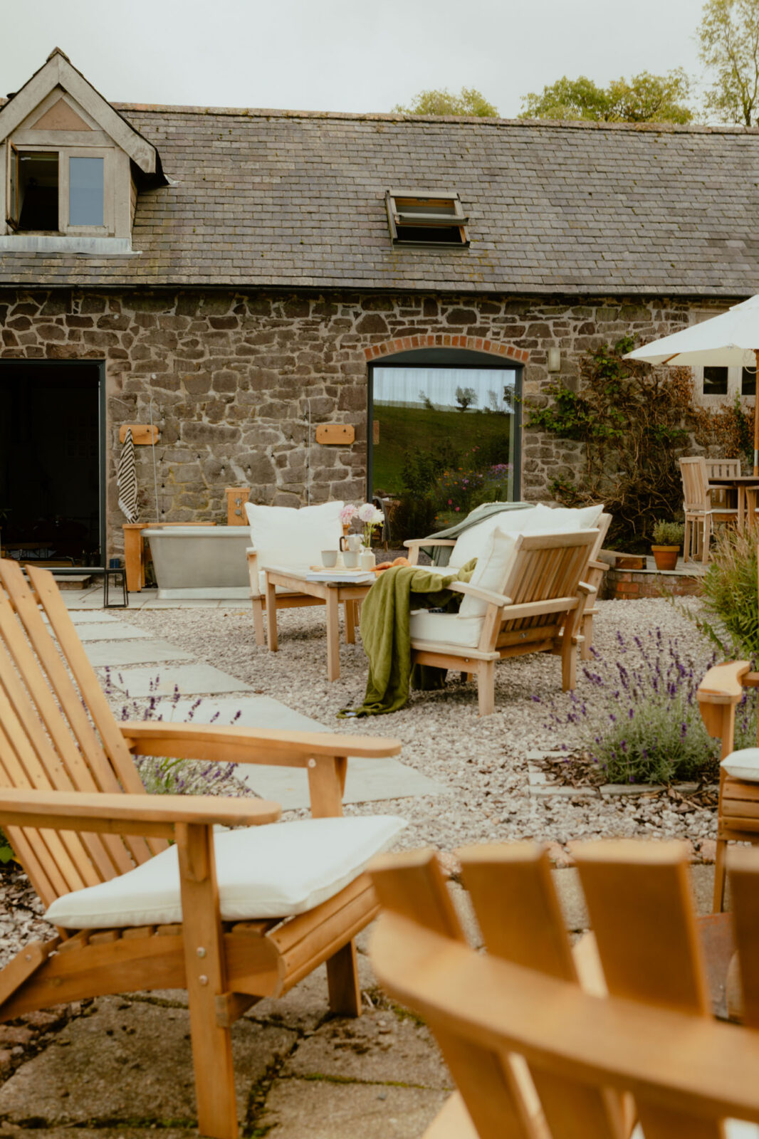 Gravel courtyard of a stone barn conversion with cushioned wooden garden sofas, a coffee table set with flowers and mugs, lavender borders, an outdoor bathtub and a parasol-shaded dining area.