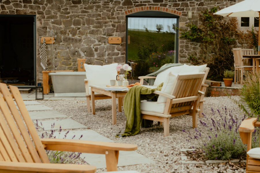 Gravel courtyard of a stone barn conversion with cushioned wooden garden sofas, a coffee table set with flowers and mugs, lavender borders, an outdoor bathtub and a parasol-shaded dining area.