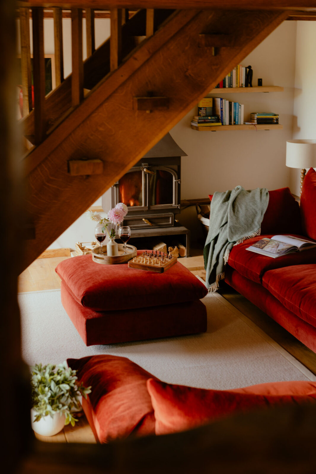 Living room seen beneath an oak staircase: a lit wood-burning stove, rust velvet sofa and footstool set with wine, dahlias and a chess board, and floating bookshelves on the wall.