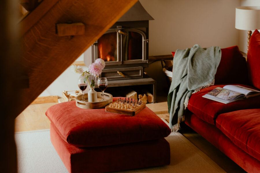 Living room seen beneath an oak staircase: a lit wood-burning stove, rust velvet sofa and footstool set with wine, dahlias and a chess board, and floating bookshelves on the wall.