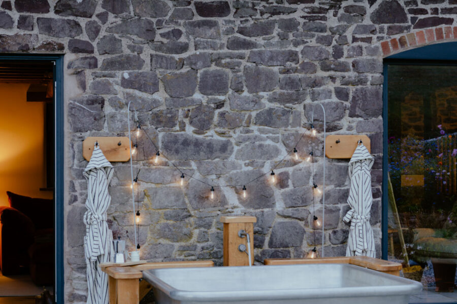 Outdoor zinc bathtub against a stone barn wall at dusk, framed by warm festoon lights and two striped robes on wooden pegs, with a glowing living room visible through the open door.