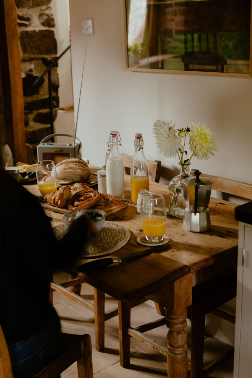 Breakfast spread on a rustic farmhouse table with croissants, pastries, sliced bread, orange juice, a bottle of milk, a moka pot and white dahlias, with a vintage radio and stone wall behind.