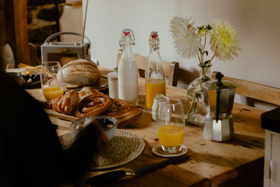 Breakfast spread on a rustic farmhouse table with croissants, pastries, sliced bread, orange juice, a bottle of milk, a moka pot and white dahlias, with a vintage radio and stone wall behind.