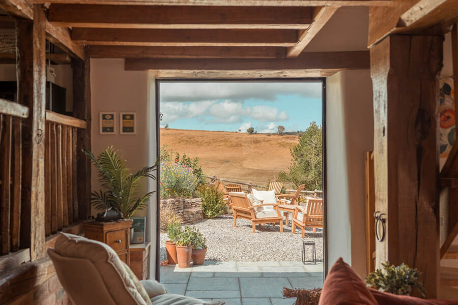 Beamed sitting room with a cream swivel chair and a rust velvet sofa, looking through a large glazed doorway to a gravel terrace with wooden lounge chairs, wildflowers, and golden hills beyond.