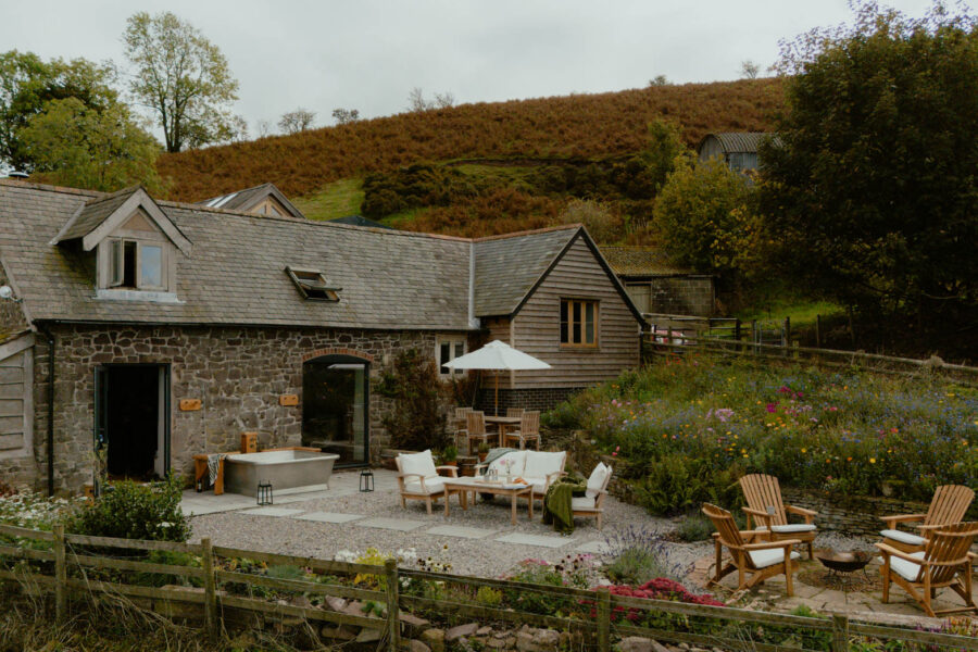 Wide view of a stone barn conversion with a slate roof and dormer window, an outdoor copper-style bath on the patio, a gravel garden with lounge seating, wildflower borders, and a circular fire pit area with wooden chairs, framed by autumn hills.