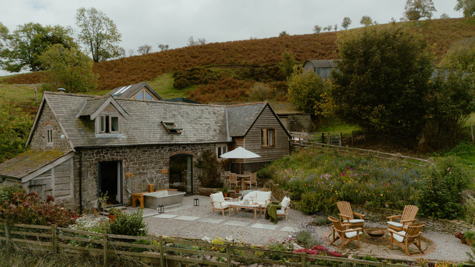 Stone and timber-clad barn conversion set below a bracken-covered hillside, with a freestanding outdoor bath by the doors, a terrace with cushioned seating and a parasol, wildflower beds, and Adirondack chairs around a fire pit.
