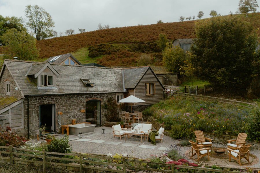 Stone and timber-clad barn conversion set below a bracken-covered hillside, with a freestanding outdoor bath by the doors, a terrace with cushioned seating and a parasol, wildflower beds, and Adirondack chairs around a fire pit.